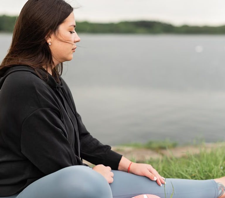 Woman meditating outdoors by a tranquil lakeside, fostering health and wellness.